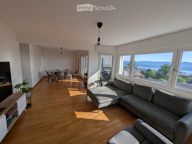 Bright living room with wooden flooring, large windows offering a scenic view, a grey sectional sofa, a dining area with a table and chairs, and a potted plant near the TV stand.
