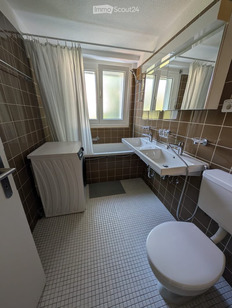 Modern bathroom with brown tiled walls, white tiled floor, double sink, toilet, bathtub, and large mirror. Features a window with frosted glass and a shower curtain.