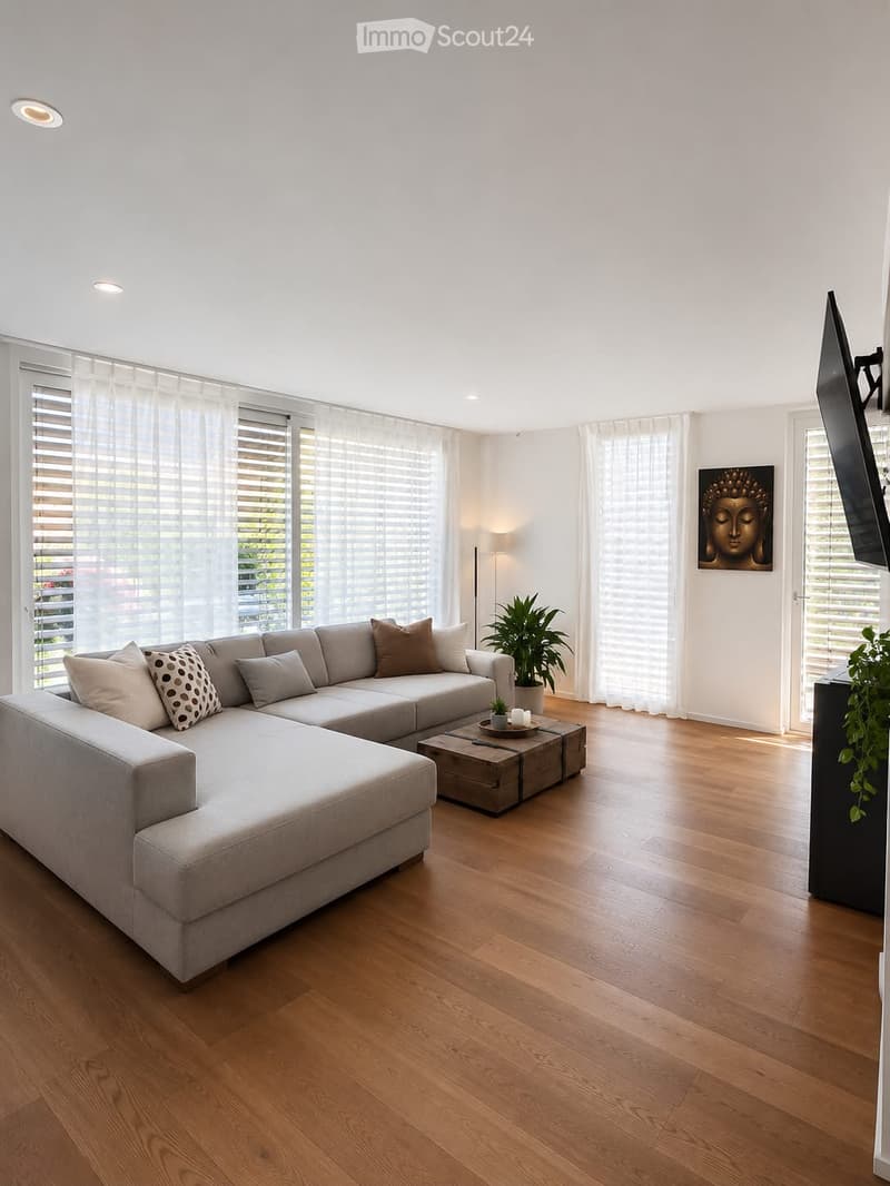 Modern living room with light wooden flooring, large windows with blinds, a grey sectional sofa, a coffee table, a floor lamp, a potted plant, and a television mounted on the wall.