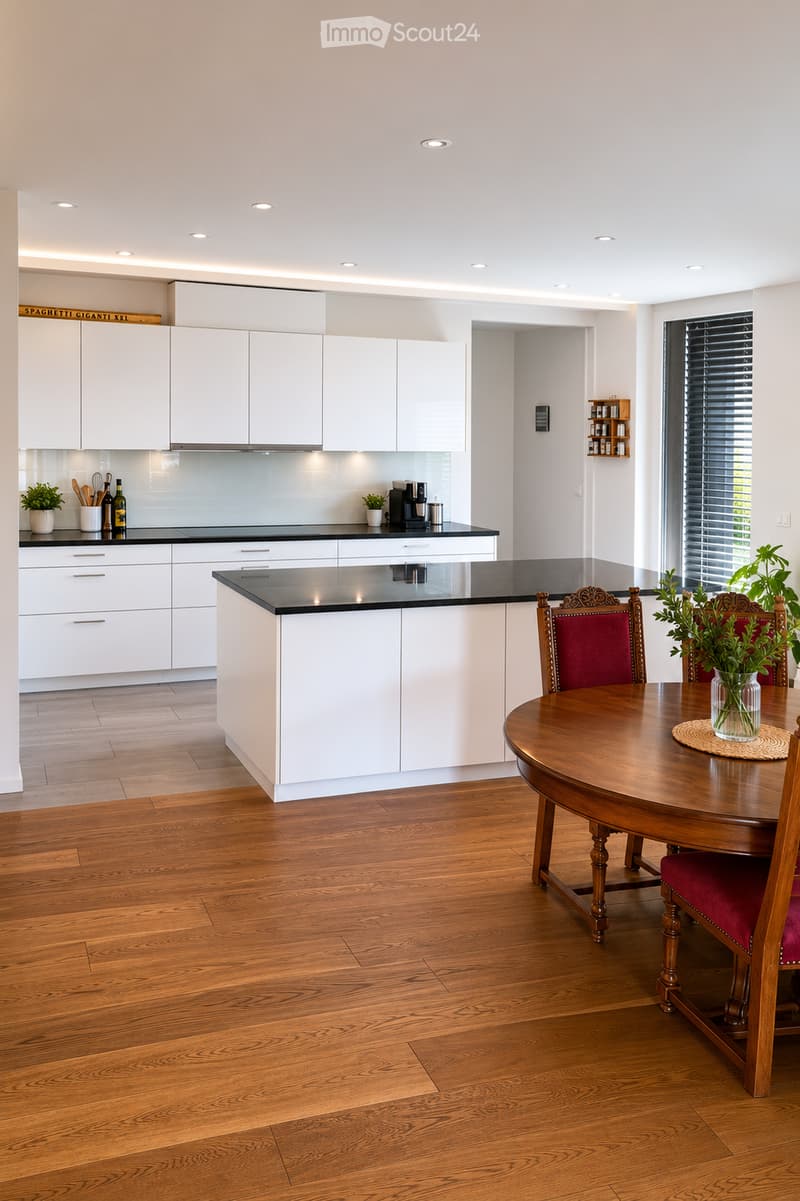 Modern kitchen with white cabinets, black countertops, and a wooden dining table with chairs. Features include recessed lighting, a coffee maker, and decorative plants.