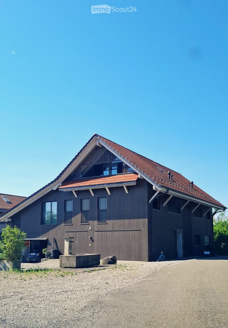 Two-story house with dark brown siding, red-tiled roof, multiple windows, and a garage. The house features a modern design with a prominent triangular roof section and a small balcony area on the upper level.