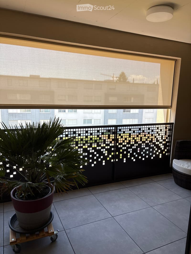 Balcony with tiled floor, large window with blinds, decorative black metal railing, potted plant on a rolling cart, and a wicker chair.
