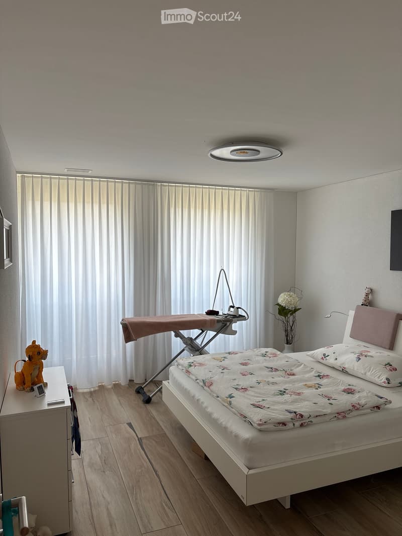 A well-lit bedroom with a white bed, floral quilt, nightstand, lamp, ironing board, vase with flowers, and decorative toys.