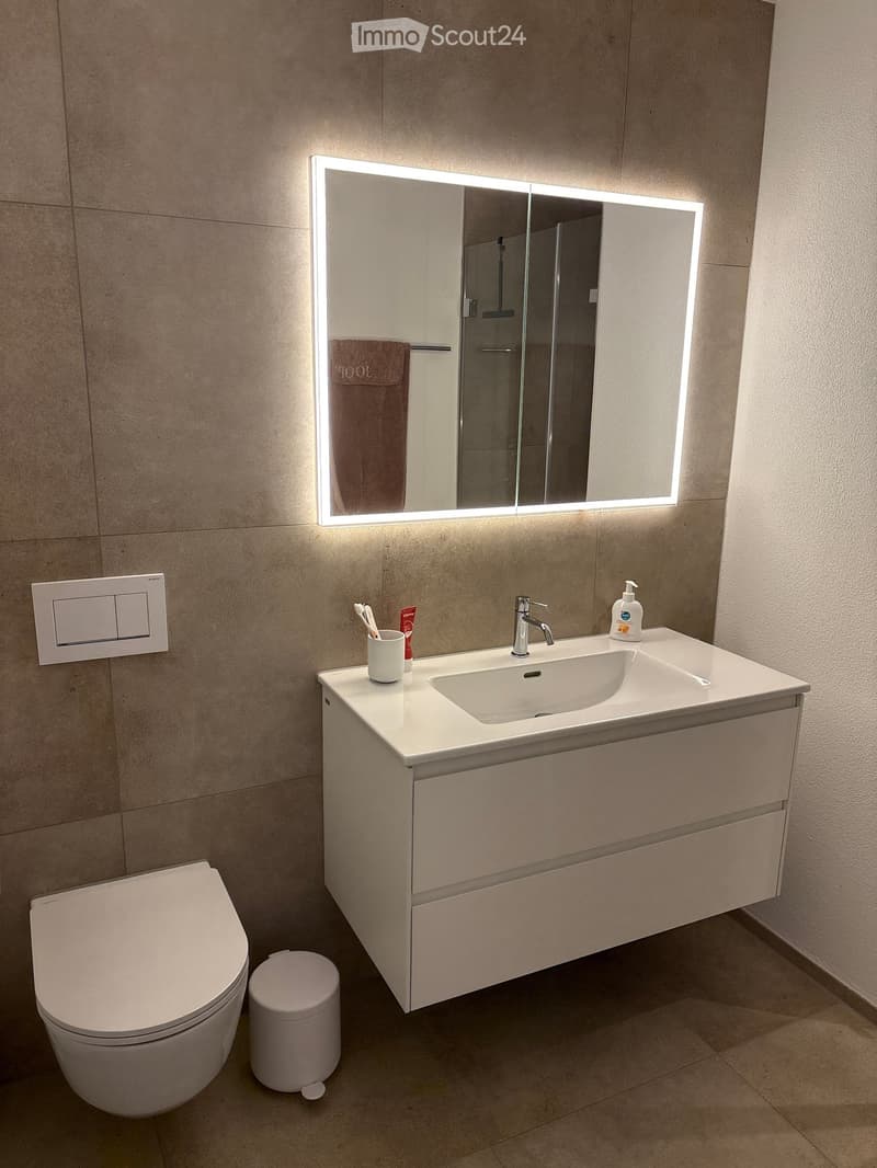 Modern bathroom with a white sink, mirror with LED lighting, toilet, and a small trash can. The sink has a faucet, a cup with toothbrushes, and a soap dispenser.