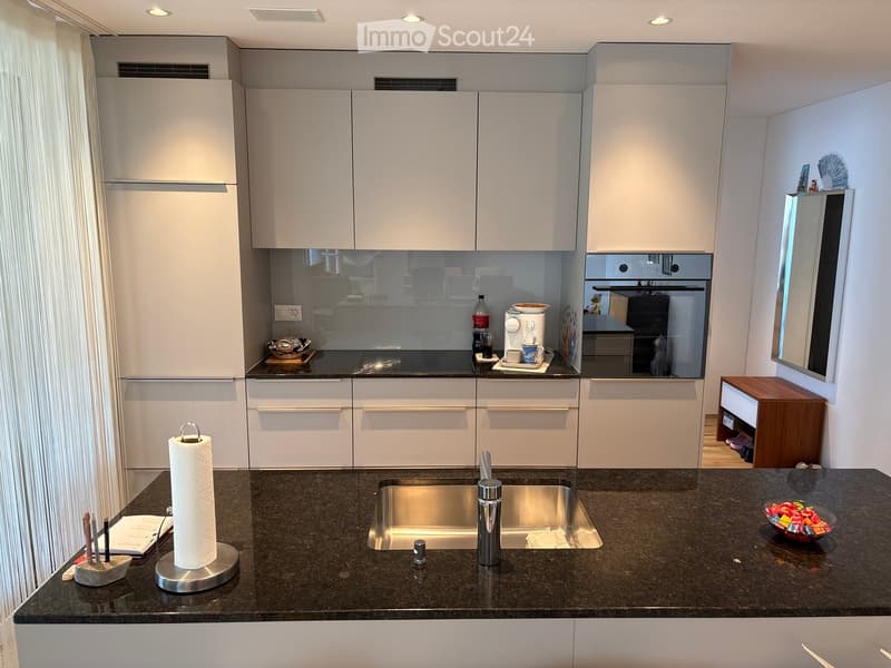 Modern kitchen with white cabinets, dark granite countertops, stainless steel sink, and various appliances including a coffee maker and oven. A small wooden table is visible in the corner.