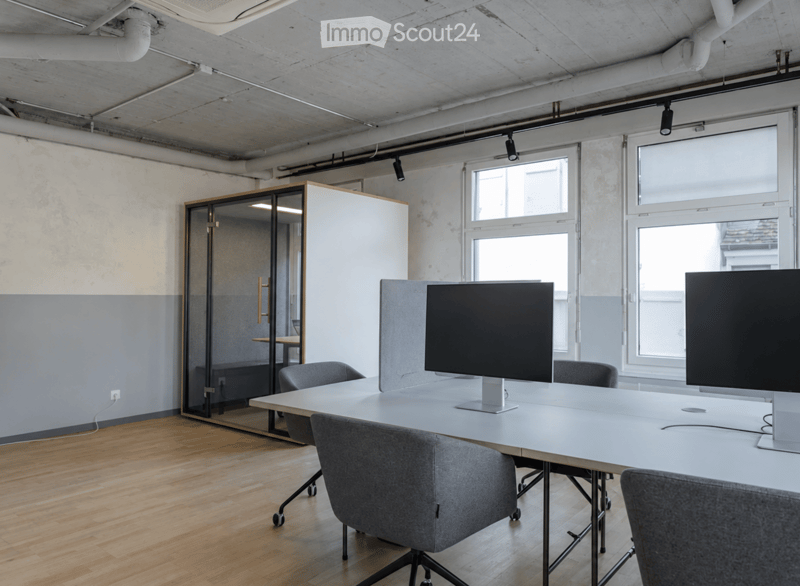 Modern office space with wooden flooring, large windows, white table, grey chairs, and two screens on the table.
