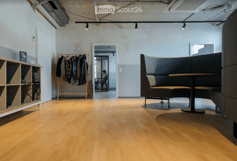 Modern, minimalist room with light wooden flooring, a large L-shaped sofa, a small round table, a wooden shelf, and a coat rack with jackets.