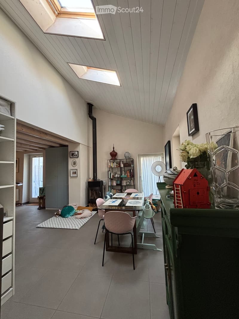 Bright, airy room with slanted ceilings and skylights, featuring a dining table with chairs, shelves, a vase with flowers, and a toy on a counter.