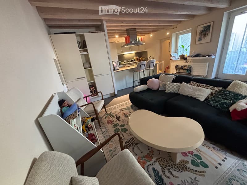 A modern living room with a dark sectional sofa, a round coffee table, and a patterned rug. The room features a kitchen area with a white island, bar stools, and modern cabinets. A cozy armchair and a small shelf with toys are also visible.