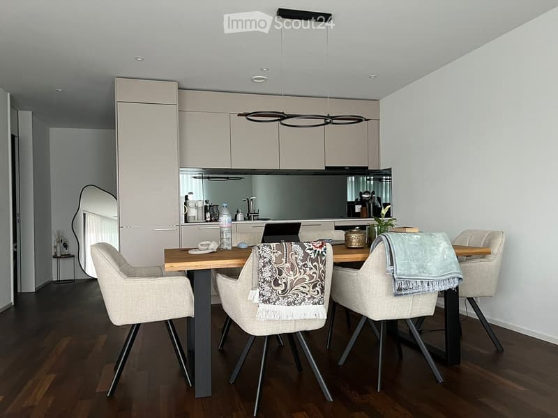 Dining room with wooden table, four chairs, modern pendant light, kitchen with appliances and mirror, wooden floor