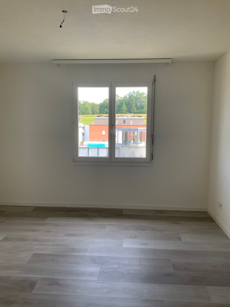 Empty room with light-colored wooden flooring and white walls, featuring a large window with a view of a green area and neighboring buildings.