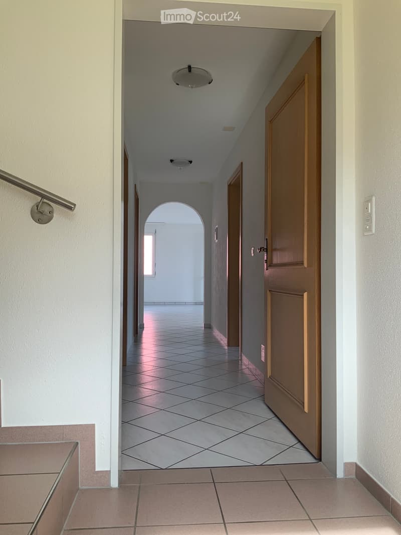 A long, tiled hallway with wooden doors on one side and a staircase railing on the other. The hallway has white walls and a ceiling light fixture.