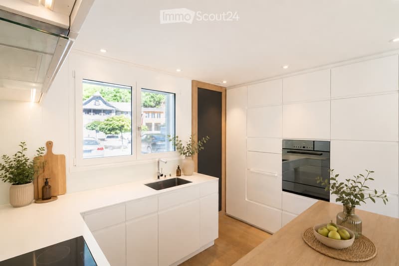 Modern kitchen with white cabinets, a central island, and a sink. Features include a built-in oven, a wooden cutting board, and decorative vases with plants.