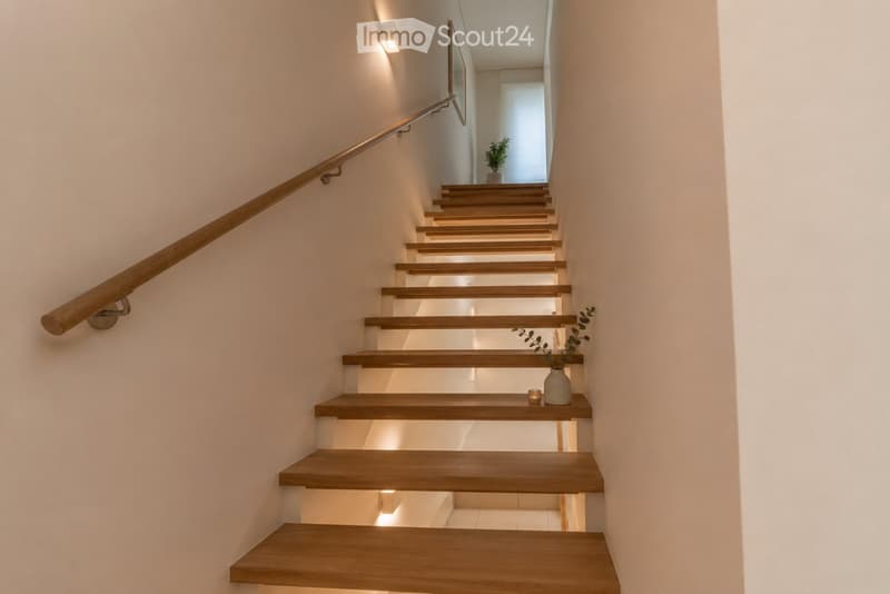 Modern staircase with wooden steps and a polished wooden handrail, illuminated by recessed lighting, with a small decorative plant and candle on a step.