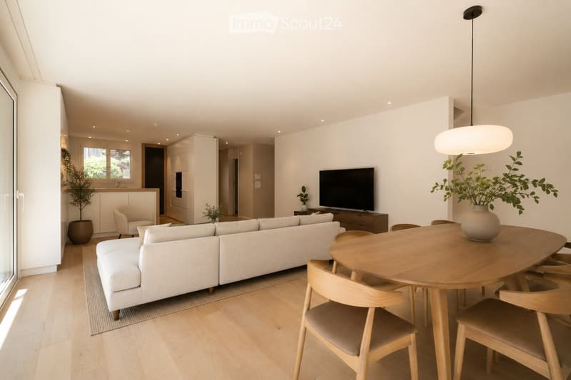 Modern living and dining area with light-colored furniture, a large sectional sofa, a round wooden dining table with chairs, a television, and decorative plants.