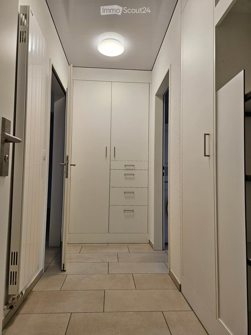 Clean, modern hallway with white walls, tiled floor, built-in storage unit with drawers, and a round ceiling light.