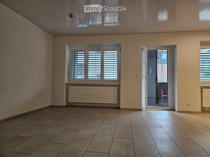 Empty room with tiled floor, white walls, large window with shutters, radiator, and a glass door leading to another space.