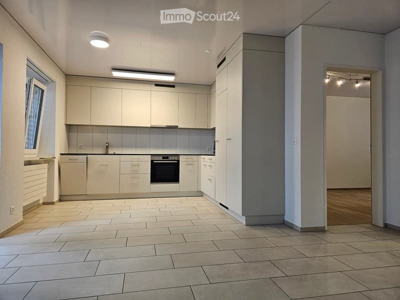 Modern, minimalist kitchen with white cabinets, a built-in oven, and a dishwasher. The space features tiled flooring and ample natural light from a window on the left.