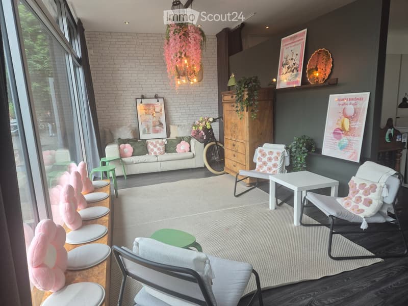 A stylish, well-lit room with a mix of modern and rustic elements. Features a white sofa with floral cushions, a hanging floral chandelier, a wooden cabinet, and several chairs and tables arranged around a central rug.