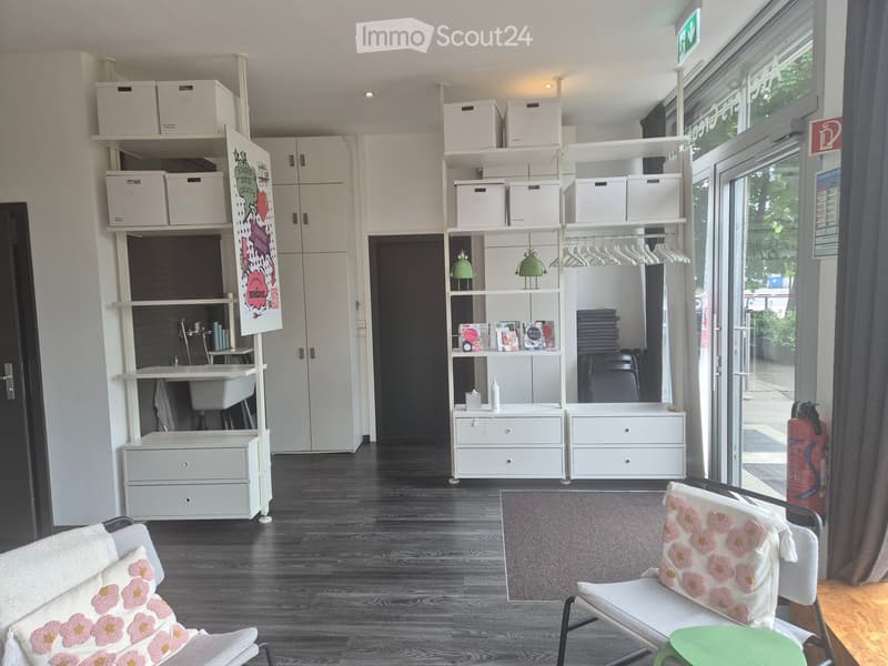 A modern, well-lit room with wooden flooring, white shelving units, and a mix of seating and storage. Features include a grey chair with a floral cushion, a small table, and a fire extinguisher near the door.