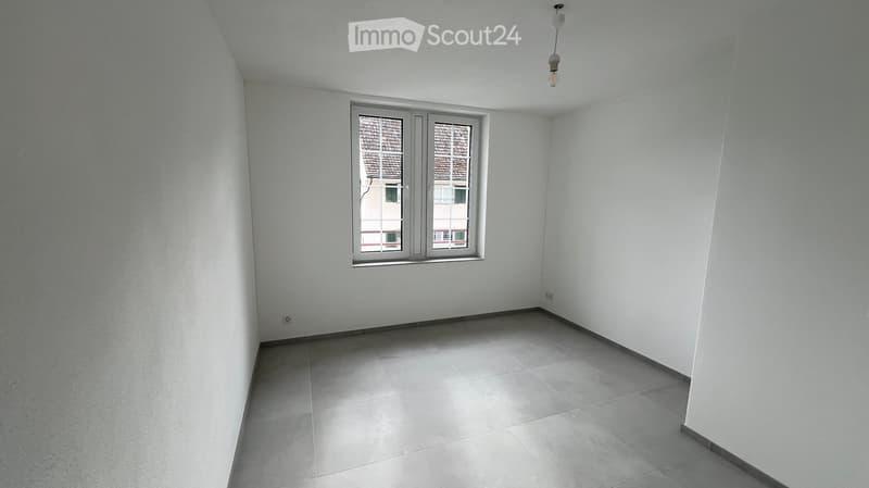 Empty room with white walls, tiled floor, and a single window with a view of a neighboring building.