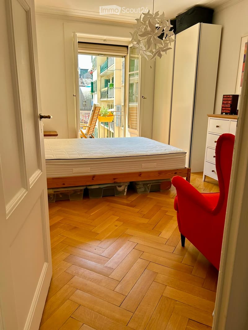 Bright bedroom with wooden flooring, a large bed, a red chair, a small table, and a view of a city street through an open door.