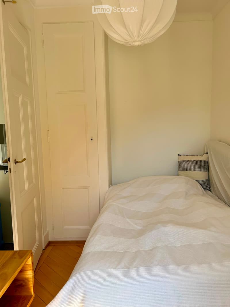 Cozy bedroom with a neatly made bed, wooden floor, and a nightstand near the door.