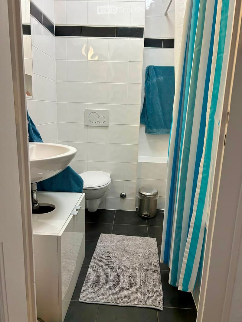 Modern bathroom with white tiles, a sink, toilet, bathtub, shower curtain, towels, and a trash can.