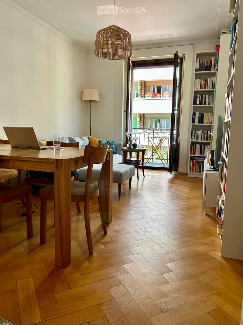 Bright, airy room with wooden flooring, a large open doorway leading to a balcony, a wooden table with a laptop, a comfortable sofa, a bookshelf filled with books, and a hanging wicker light fixture.