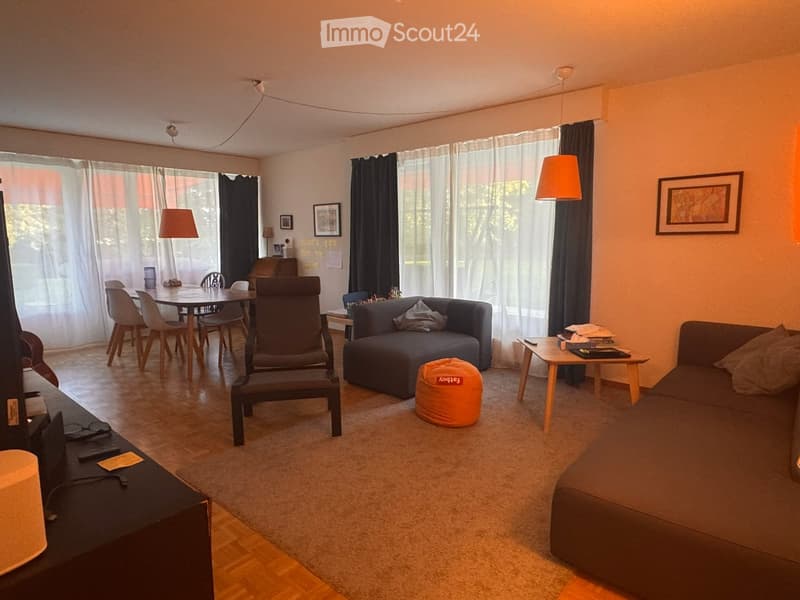A modern living room with a sofa, a chair, a small table, a round stool, a dining table with chairs, and a TV stand with a screen. The room has large windows with curtains and is well-lit with hanging lamps.