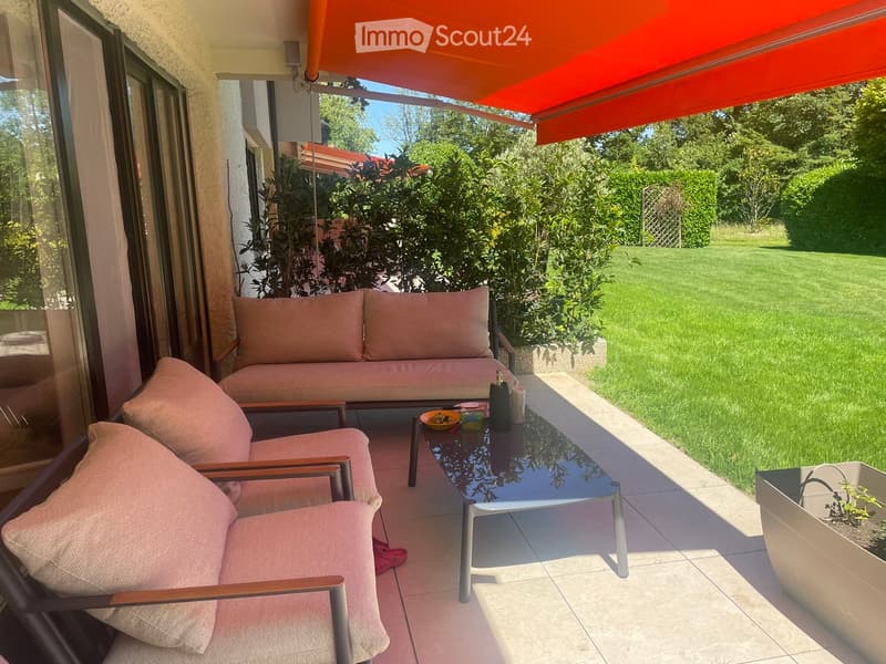 Balcony with a sofa, two chairs, a coffee table, and a bottle on the table, surrounded by greenery and a lawn.