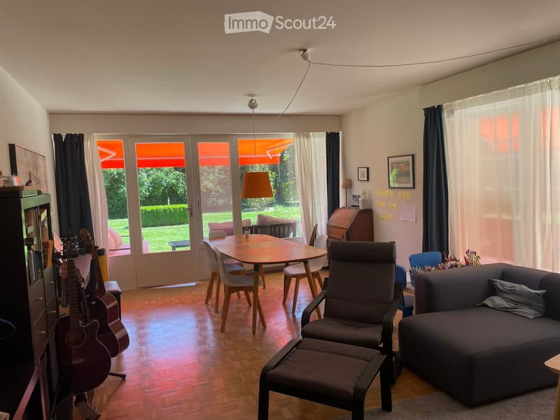 A modern living room with a dark grey sofa, a wooden dining table with chairs, a guitar, and a comfortable chair with an ottoman. Large windows with curtains lead to a garden view.