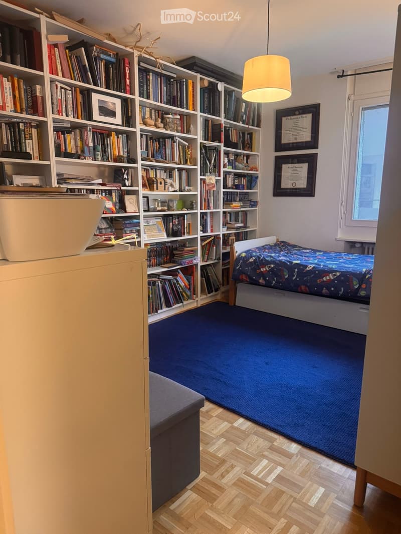Cozy bedroom with a single bed, large bookshelf filled with books, a laptop on a cabinet, and a hanging lamp.