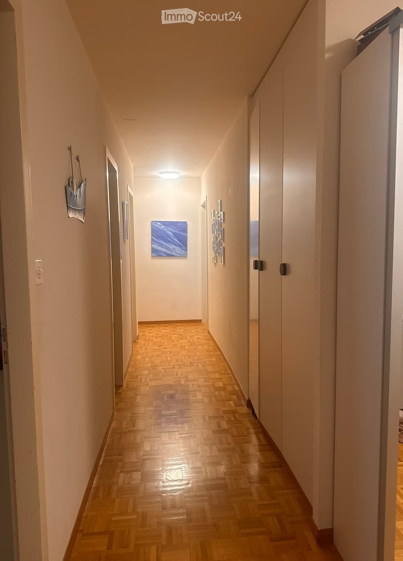 A long, narrow hallway with wooden herringbone flooring, white walls, and recessed ceiling lighting. Features several doors and a small artwork on the wall.