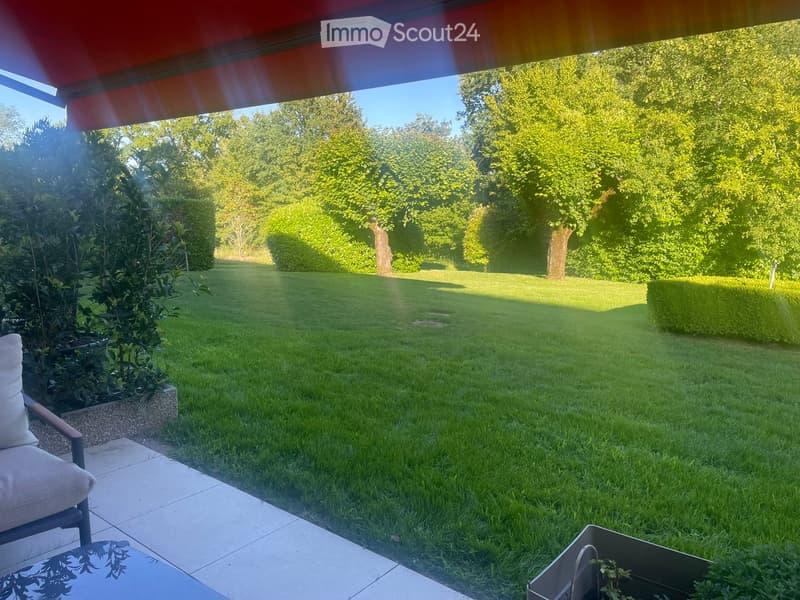 Well-maintained garden with a neatly trimmed lawn, mature trees, and shrubs. A patio area with a cushioned chair and small table is visible on the left side.