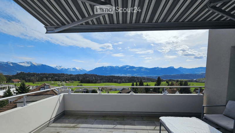 Balcony with a retractable awning, overlooking a scenic valley and mountain range, with a few lounge chairs placed on the tiled floor.