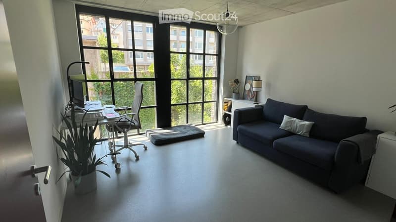 Modern living room with large windows, a dark blue sofa, a white desk with a computer, a chair, and a potted plant.