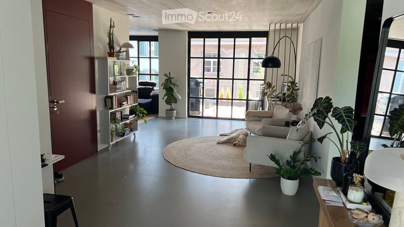 Modern living room with large windows, white walls, grey sofa, white armchair, round rug, floor lamp, bookshelf, and potted plants.