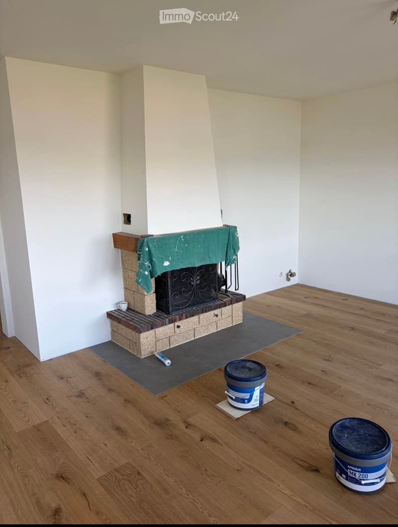 Empty room with wooden flooring, white walls, and a fireplace with a green cloth draped over it. There are two paint buckets and a paint roller on the floor.