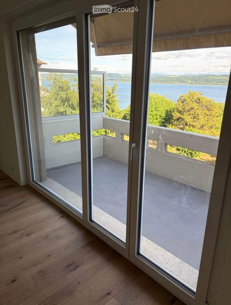 View of a lake and surrounding greenery through large sliding glass doors leading to a balcony with wooden railings.