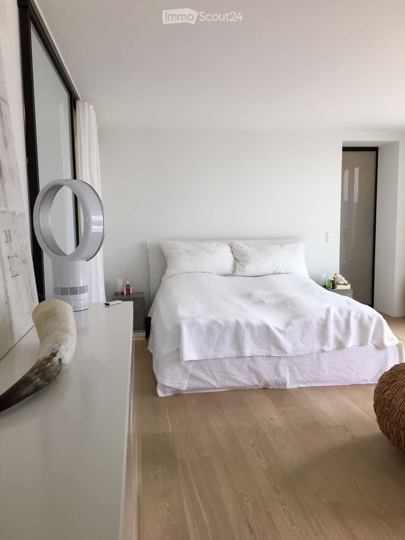 Modern bedroom with a large bed, nightstand, mirror, fan, and a small table with a decorative item on it.