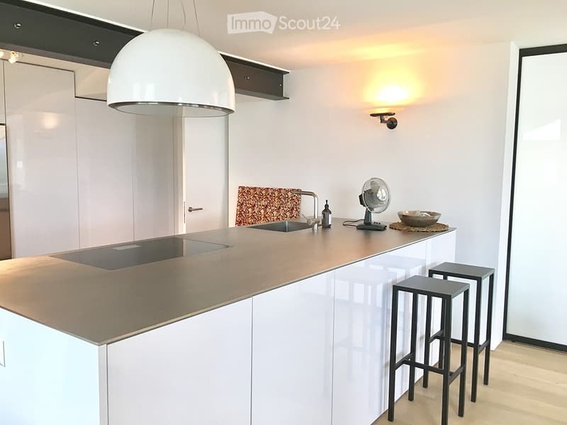 Modern kitchen with white cabinets, a central island, and a built-in stove. Features include a hanging pendant light, a wall-mounted fan, and two stools at the island.