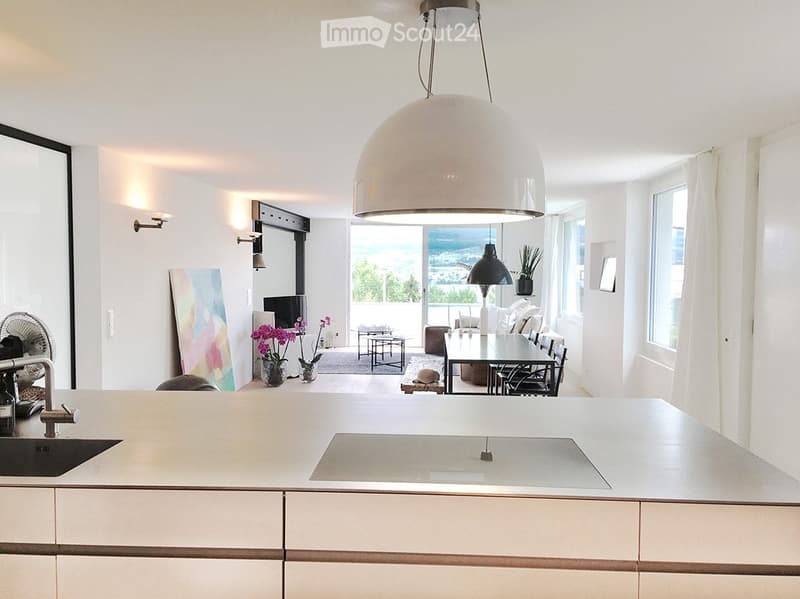 Modern kitchen with white cabinets, a large island, and a view of the ocean through large windows. The space includes a dining area with a table and chairs, a fan, and decorative vases with flowers.