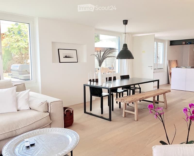 Bright, open living and dining area with light wood flooring, white walls, and large windows. Features a beige sofa, glass coffee table, black dining table with chairs, wooden bench, and decorative vases.