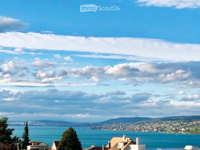 Scenic view of a lake with surrounding hills and a town in the distance under a partly cloudy sky.