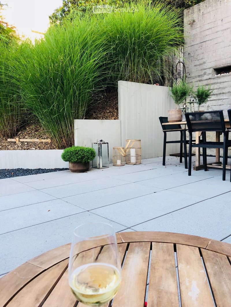 Modern outdoor patio with white tiled flooring, wooden furniture, potted plants, and decorative lanterns.