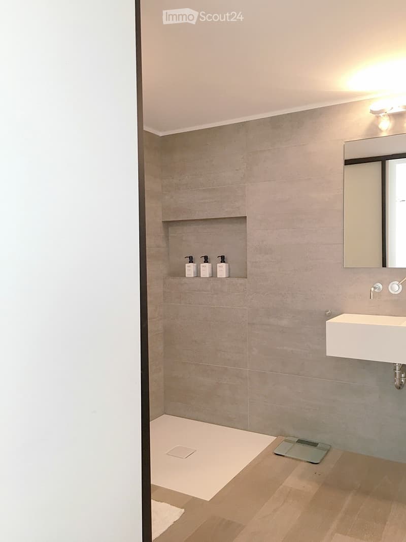Modern bathroom with a white sink, mirror, and a shelf holding four bottles. The floor is light wood, and the walls are tiled in a neutral tone.