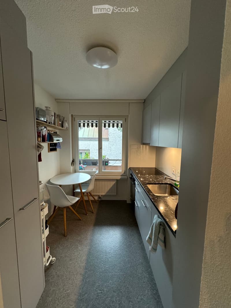 Compact kitchen with a round white table, two white chairs, a sink, and built-in cabinets.