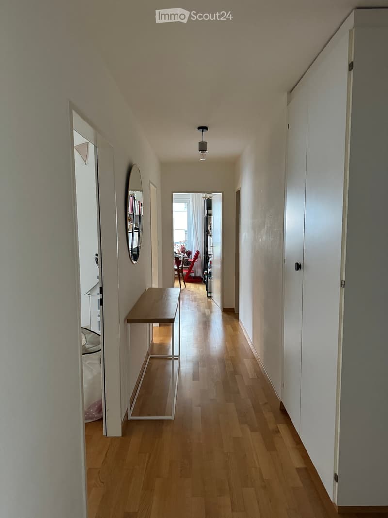 A narrow hallway with wooden flooring, white walls, and a small wooden console table on the left side. A mirror is mounted on the wall, and a doorway leads to another room with visible furniture.