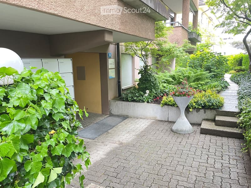 Garden area with paved walkway, greenery, and a modern building entrance.